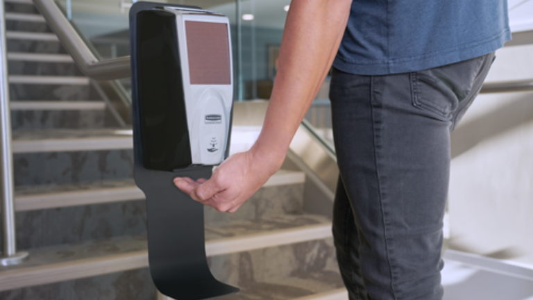Person using a touchless hand sanitizer dispenser near stairs in a modern building.
