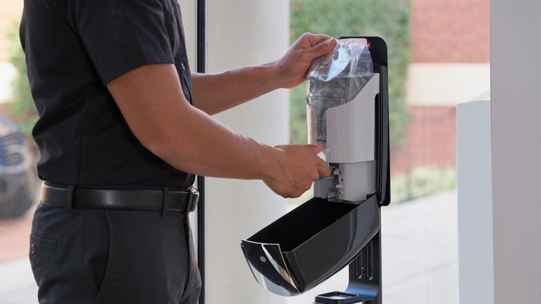 Person refilling automatic hand sanitizer dispenser with a transparent sanitizer bag inside a building.