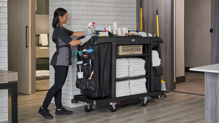 Hotel housekeeper organizing cleaning cart with towels and supplies in modern hallway.